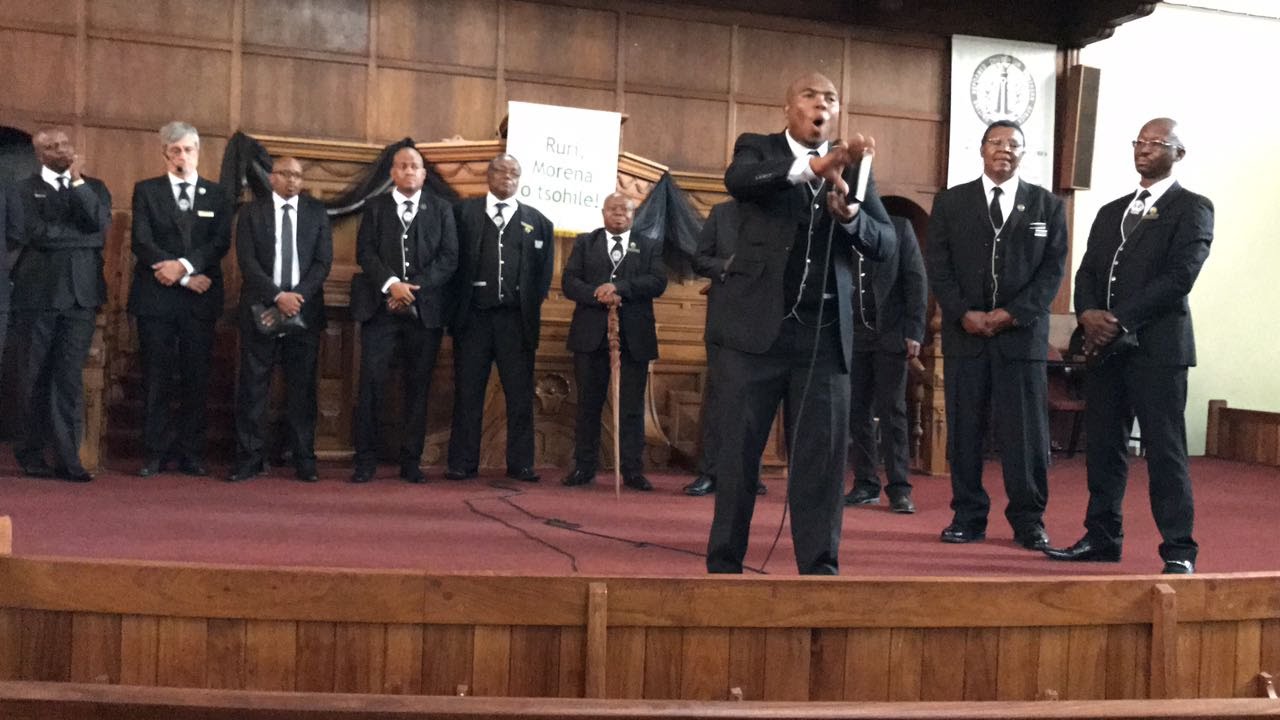 Image of 11 Men in URCSA CMM uniform - a white shirt, black suit and black long pants.
The mend are standing in front of the church podium that is decorated with a cloth written
'Ruri Moren o tsohile'. One of the men is standing in front of other and is talking on a microphone.