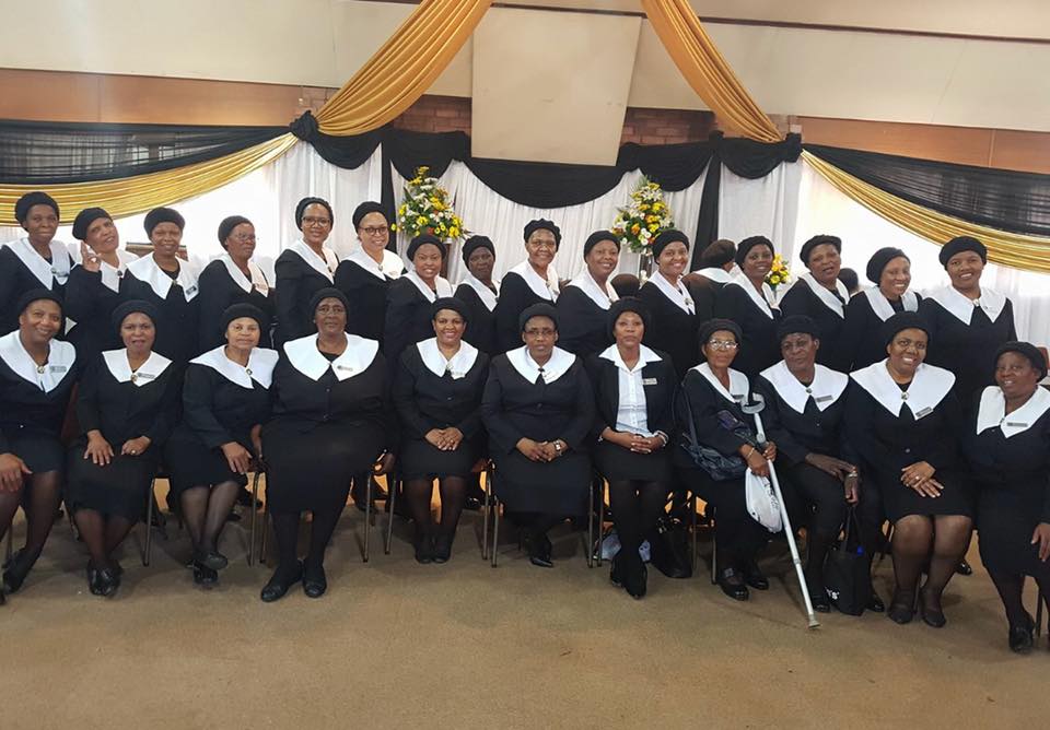 Women wearini URCSA CWM uniform
posing in two rows. The first row consisist 11 women sitting and the second row row consists of 15 women
standing. On the background there is decoration similar to tent decor.