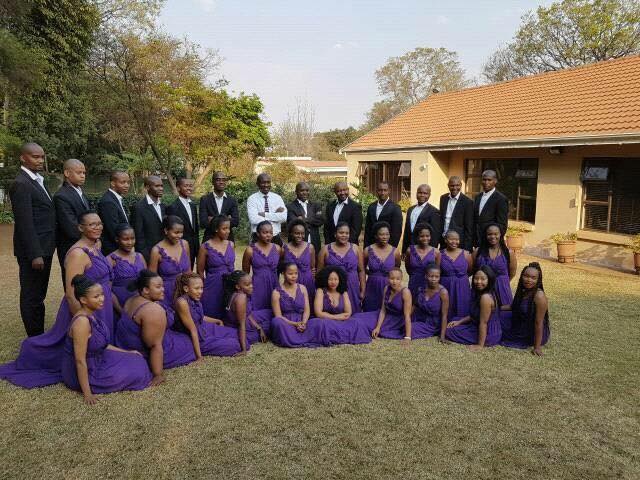 Melodi ya Tshwane choir members posing at a garden in three rows. On the first row
are women wearing purple dresses sitting on the grass of the garden. On the second
row are women kneeling on the garden grass wearing purple dresses. On the third row
are men wearing black suits and white shirts, standing. On the background on the left
there is a townhouse and on the right there are trees.