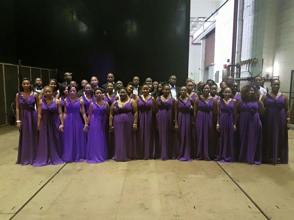 Melodi ya Tshwane choir members posing at a backstage in three rows. On the first and
second rows are women wearing purple dresses standing. On the third row are men wearing
black suits and white shirts also standing.